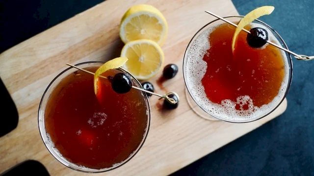 Two cocktails garnished with cherries and lemon twists are on a wooden board, accompanied by lemon slices and extra cherries nearby.